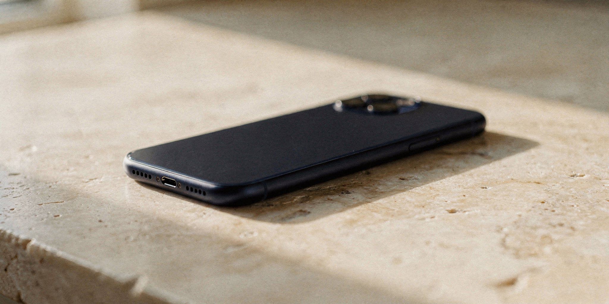 A single modern smartphone resting face-up on a warm travertine surface in soft afternoon window light